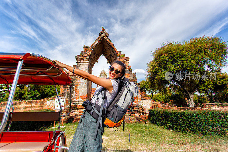 年轻的亚洲女性旅行者背包旅行站在出租车或Tuk Tuk和快乐的乐趣与老寺庙(Wat Mahathat)背景，大城府，泰国
